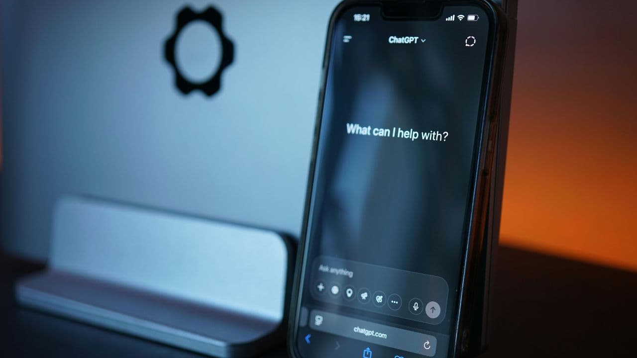 Phone screen displays ChatGPT prompt 'What can I help with?' on a desk, with a blurred gear icon in the background at left.