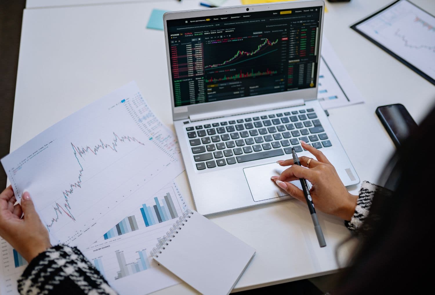 Person using a laptop with stock market charts on screen while reviewing printed graphs on a white desk
